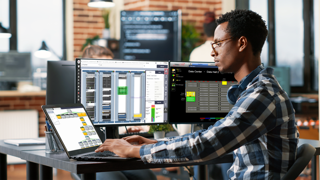 A man working on a laptop in an office. In the background there are two additional monitors. On one is a DCIM rack view. On the other is a data center visualization in a BAS system.