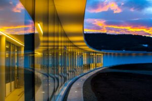 a curved glass wall lit by yellow lights underneath a cloudy sky