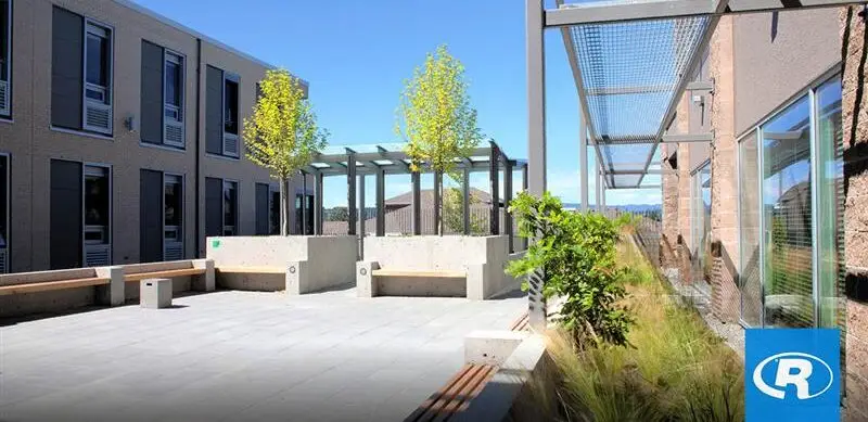 A landscaped outdoor courtyard between two modern building wings, featuring concrete seating, young trees, and metal architectural shading elements under a clear blue sky.