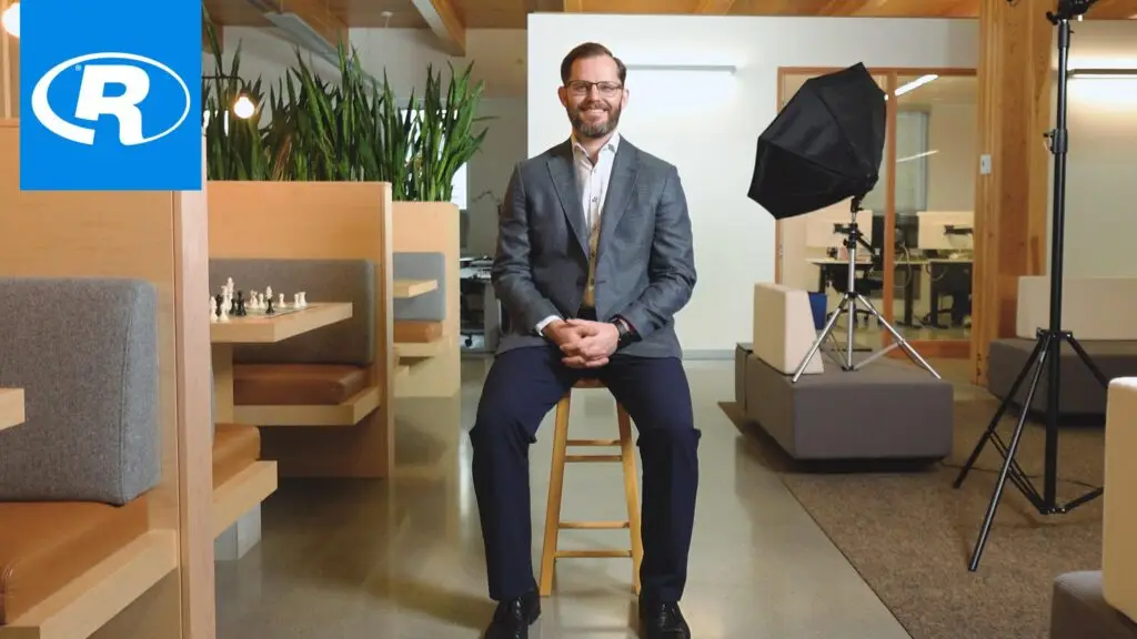 Man seated for interview in office with lighting setup, plants, and Reliable Controls logo in corner.